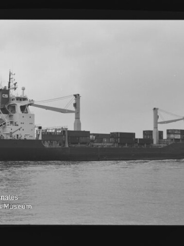 Schwarz-weiß-Seitenansicht eines Frachtschiffs mit zwei Ladekränen und gestapelten Containern auf ruhigem Wasser unter bedecktem Himmel; Bild zeigt Filmrand mit AGFA APX 100-Markierung und Wasserzeichen des Internationalen Maritimen Museums Hamburg.