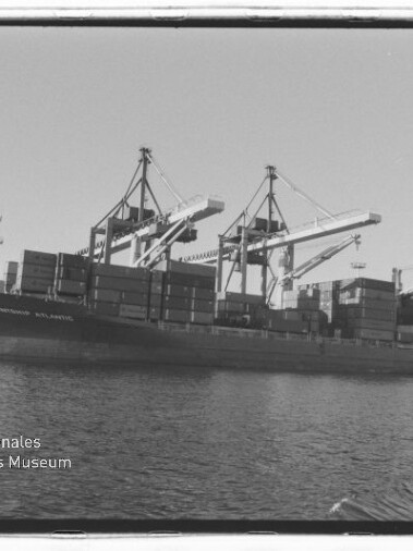 Schwarzweiß-Foto eines mit Containern beladenen Frachtschiffs am Hafenpier, davor Wasser und mehrere große Containerkräne sowie Hafeninfrastruktur; Wasserzeichen des Internationalen Maritimen Museums Hamburg in der Ecke.