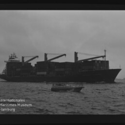 Beladenes Containerschiff mit drei Ladekränen und gestapelten Containern vor trübem Himmel, im Vordergrund ein kleines Motorboot auf dem Meer; unten links das Logo des Internationalen Maritimen Museums Hamburg.