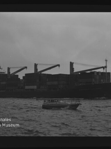 Beladenes Containerschiff mit drei Ladekränen und gestapelten Containern vor trübem Himmel, im Vordergrund ein kleines Motorboot auf dem Meer; unten links das Logo des Internationalen Maritimen Museums Hamburg.