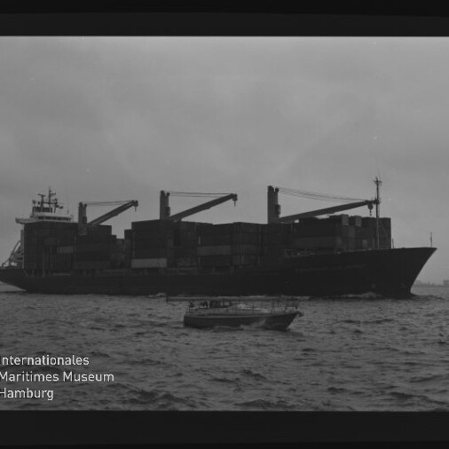 Beladenes Containerschiff mit drei Ladekränen und gestapelten Containern vor trübem Himmel, im Vordergrund ein kleines Motorboot auf dem Meer; unten links das Logo des Internationalen Maritimen Museums Hamburg.