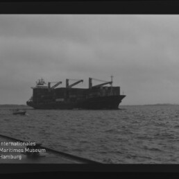 Schwarzweißaufnahme eines beladenen Frachtschiffs mit Containerkränen auf leicht welliger See unter bewölktem Himmel; links unten das Logo des Internationalen Maritimen Museums Hamburg und am Bildrand die Filmaufschrift AGFA APX 100 sichtbar.
