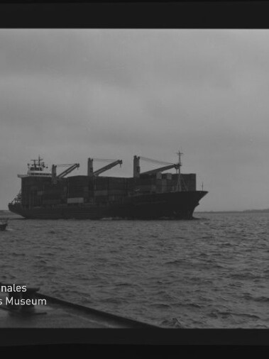Schwarzweißaufnahme eines beladenen Frachtschiffs mit Containerkränen auf leicht welliger See unter bewölktem Himmel; links unten das Logo des Internationalen Maritimen Museums Hamburg und am Bildrand die Filmaufschrift AGFA APX 100 sichtbar.