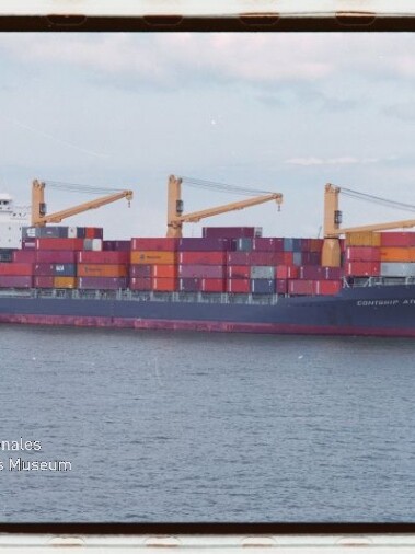 Containerschiff mit gestapelten bunten Containern und drei gelben Ladekränen fährt auf ruhiger See unter hellem Himmel; unten links sichtbares Logo des Internationalen Maritimen Museums Hamburg.