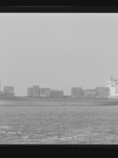Schwarz-weiß-Foto eines Containerschiffs in Seitenansicht auf ruhiger See mit übereinandergestapelten Ladecontainern, leicht diesigem Horizont, sichtbarem Filmrand und der Aufschrift AGFA APX 100 sowie Museumslogo unten links