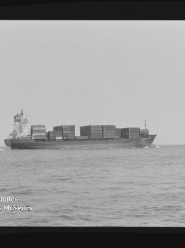 Schwarzweiß-Seitenansicht eines beladenen Containerschiffs mit übereinander gestapelten Containern, das auf ruhiger See nach rechts fährt; schwarzer Bildrand und Wasserzeichen des Internationalen Maritimen Museums Hamburg links unten.