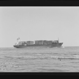 Schwarzweißaufnahme eines beladenen Containerschiffs auf ruhiger See, seitliche Ansicht mit gestapelten Containern sowie sichtbarem Negativrahmen und Wasserzeichen des Internationalen Maritimen Museums Hamburg