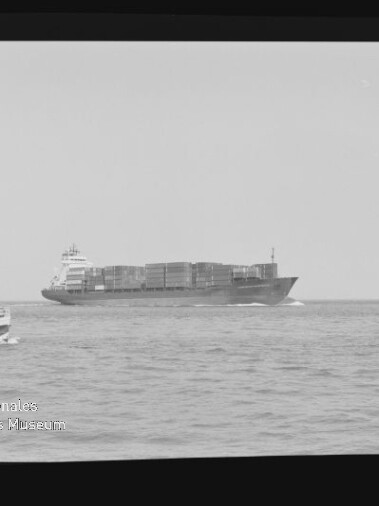 Schwarzweißaufnahme eines großen Containerschiffs auf offenem Meer, links vorn ein kleines Ausflugsboot, umgeben von Filmrand mit Schriftzug AGFA APX 100 und Museumslogo.