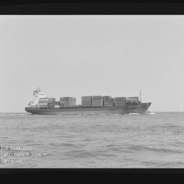 Schwarzweiß-Foto eines beladenen Containerschiffs in Seitenansicht mit aufgetürmten Containerblöcken, das auf ruhiger See fährt und Bug- sowie Heckwellen zeigt; schwarzer Filmrand mit Nummerierung und Wasserzeichen des Internationalen Maritimen Museums Hamburg sind sichtbar.
