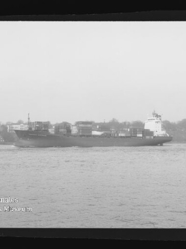 Schwarzweiß-Aufnahme eines Containerschiffs mit aufgestapelten Containern, das auf ruhigem Wasser vor einem Ufer mit Gebäuden und Bäumen fährt; schwarzer Filmrand und Museumslogo unten links sichtbar.
