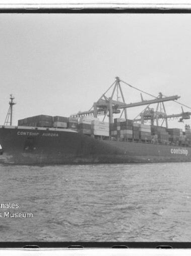 Schwarzweißaufnahme des Containerschiffs Contship Aurora am Kai, beladen mit gestapelten Containern und umgeben von großen Containerkränen, mit Wasser im Vordergrund und Museumswasserzeichen unten links.