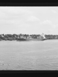 Schwarzweißaufnahme eines Containerschiffs mit gestapelten Ladecontainern auf ruhigem Wasser, vor einem baumbestandenen Ufer mit vereinzelten Häusern; Filmrand und Museums-Wasserzeichen unten links sichtbar.