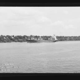 Schwarzweißaufnahme eines Containerschiffs mit gestapelten Ladecontainern auf ruhigem Wasser, vor einem baumbestandenen Ufer mit vereinzelten Häusern; Filmrand und Museums-Wasserzeichen unten links sichtbar.