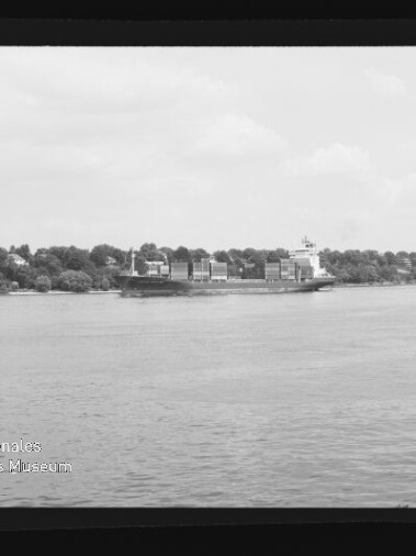 Schwarzweißaufnahme eines Containerschiffs mit gestapelten Ladecontainern auf ruhigem Wasser, vor einem baumbestandenen Ufer mit vereinzelten Häusern; Filmrand und Museums-Wasserzeichen unten links sichtbar.