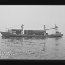 Schwarz-weiß-Seitenansicht eines Containerfrachtschiffs mit zwei Ladekränen und gestapelten Containern, das ruhig auf dem Meer liegt; Wasserzeichen des Internationalen Maritimen Museums Hamburg unten links.