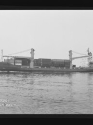 Schwarzweiß-Foto eines Containerschiffs mit zwei Ladekränen und gestapelten Containern, das ruhig auf dem Meer fährt; im Bildrand sichtbare Filmrahmung mit Aufdruck AGFA PAN 100 und Wasserzeichen des Internationalen Maritimen Museums Hamburg.