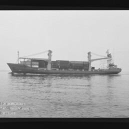 Schwarzweiß-Foto eines Containerschiffs mit zwei Ladekränen und gestapelten Containern, das ruhig auf dem Meer fährt; im Bildrand sichtbare Filmrahmung mit Aufdruck AGFA PAN 100 und Wasserzeichen des Internationalen Maritimen Museums Hamburg.