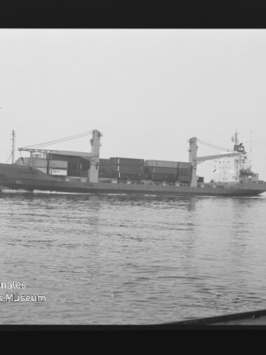 Schwarzweißaufnahme eines seitenansichtigen Containerschiffs mit zwei Ladekränen und gestapelten Containern auf ruhigem Wasser; Filmrand und Museumslogo unten links sichtbar.