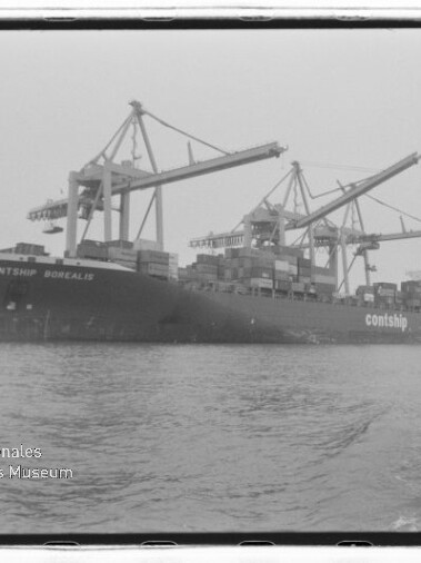 Schwarzweißaufnahme eines großen Containerschiffs (Contship Borealis) am Kai, von mehreren Hafenkranen beladen, mit aufgetürmten Containern auf dem Deck und ruhigem Hafenwasser im Vordergrund; links unten Wasserzeichen des Internationalen Maritimen Museums Hamburg.