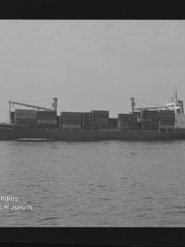 Schwarzweiß-Seitenansicht eines Containerschiffs mit gestapelten Containerblöcken und zwei Ladekränen, das auf ruhiger See vor flachem Horizont fährt; unten links sichtbar das Logo des Internationalen Maritimen Museums Hamburg.