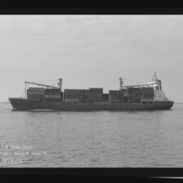 Schwarz-weiß-Foto eines in Seitenansicht fahrenden Containerschiffs auf ruhiger See mit übereinandergestapelten Containern und zwei Ladekränen auf dem Deck, im Vordergrund Wasserfläche und unten links das Logo des Internationalen Maritimen Museums Hamburg