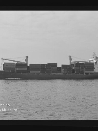 Schwarz-weiß-Foto eines in Seitenansicht fahrenden Containerschiffs auf ruhiger See mit übereinandergestapelten Containern und zwei Ladekränen auf dem Deck, im Vordergrund Wasserfläche und unten links das Logo des Internationalen Maritimen Museums Hamburg
