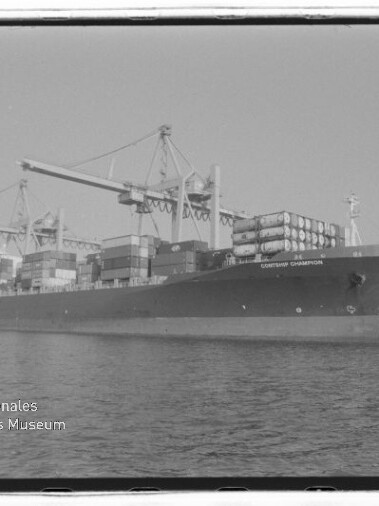 Schwarzweiß-Foto eines großen Containerschiffs am Kai mit übereinander gestapelten Containern und mehreren Hafenkränen, ruhigem Wasser im Vordergrund und einem Wasserzeichen des Internationalen Maritimen Museums Hamburg unten links.