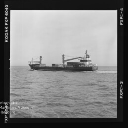 Schwarzweißaufnahme eines Frachtschiffs mit zwei Ladekränen und gestapelten Containern auf ruhiger See; Negativfilmrand mit der Aufschrift KODAK FXP und Wasserzeichen des Internationalen Maritimen Museums Hamburg sichtbar.
