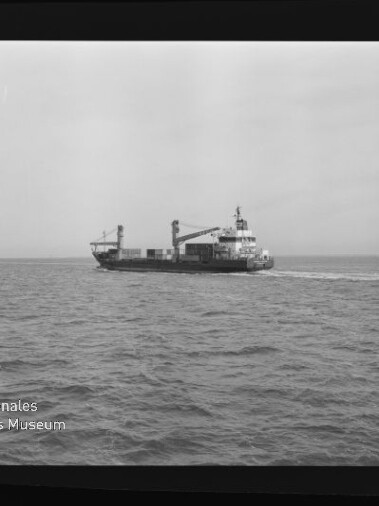 Schwarzweiß-Filmbild mit sichtbarem Kodak-Filmrand und Museumswasserzeichen, das ein mittleres Frachtschiff mit Ladekränen auf ruhiger See vor flachem Horizont zeigt.