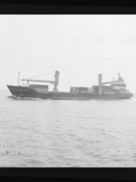 Schwarzweiß-Fotografie eines Frachtschiffs in Seitenansicht mit zwei Ladekränen und mehreren Containern an Deck, fahrend auf ruhiger See, umgeben von einem Filmnegativrand mit der Aufschrift ‚KODAK FXP 6040‘ und Wasserzeichen des Internationalen Maritimen Museums Hamburg.