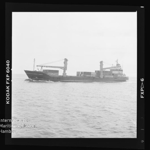 Schwarzweiß-Fotografie eines Frachtschiffs in Seitenansicht mit zwei Ladekränen und mehreren Containern an Deck, fahrend auf ruhiger See, umgeben von einem Filmnegativrand mit der Aufschrift ‚KODAK FXP 6040‘ und Wasserzeichen des Internationalen Maritimen Museums Hamburg.