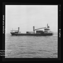 Schwarzweiß-Aufnahme eines mittleren Containerschiffs mit zwei Deckkränen und mehreren aufgestapelten Containern, seitlich fahrend auf ruhiger See; Filmrand mit 