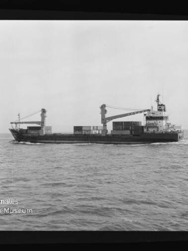 Schwarzweiß-Aufnahme eines mittleren Containerschiffs mit zwei Deckkränen und mehreren aufgestapelten Containern, seitlich fahrend auf ruhiger See; Filmrand mit 