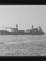 Schwarzweiß-Foto eines Containerschiffs in Seitenansicht auf ruhiger See mit aufgestapelten Containern und Ladekränen, umgeben von sichtbarem Filmrahmenrand und Wasserzeichen des Internationalen Maritimen Museums Hamburg.