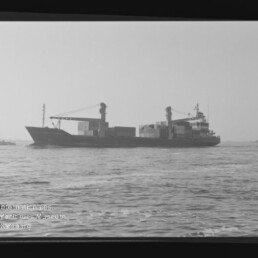Schwarzweiß-Foto eines Containerschiffs in Seitenansicht auf ruhiger See mit aufgestapelten Containern und Ladekränen, umgeben von sichtbarem Filmrahmenrand und Wasserzeichen des Internationalen Maritimen Museums Hamburg.