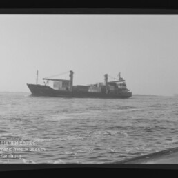 Schwarzweißaufnahme eines Containerschiffs mit aufgestapelten Containern und Ladekränen, das bei ruhiger See am Horizont fährt; im Vordergrund welliges Wasser, links unten Museumslogo und sichtbare Filmränder.
