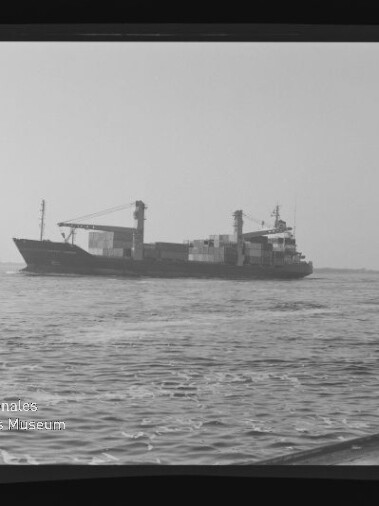 Schwarzweißaufnahme eines Containerschiffs mit aufgestapelten Containern und Ladekränen, das bei ruhiger See am Horizont fährt; im Vordergrund welliges Wasser, links unten Museumslogo und sichtbare Filmränder.