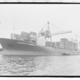Containerschiff CONTSHIP EUROPE am Industriehafen, beladen mit gestapelten Containern und von großen Umschlagkränen umgeben, Wasser im Vordergrund.