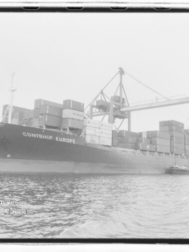 Containerschiff CONTSHIP EUROPE am Industriehafen, beladen mit gestapelten Containern und von großen Umschlagkränen umgeben, Wasser im Vordergrund.