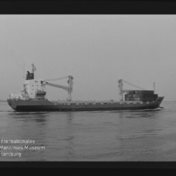 Schwarzweiß-Foto eines Frachtschiffs längsseits auf ruhigem Wasser mit zwei Deckskranen und gestapelten Containern am Heck; linke untere Ecke zeigt das Logo des Internationalen Maritimen Museums Hamburg.