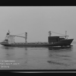Frachtschiff mit zwei Deckkränen und gestapelten Containern fährt auf ruhigem Wasser, links unten das Logo des Internationalen Maritimen Museums Hamburg.