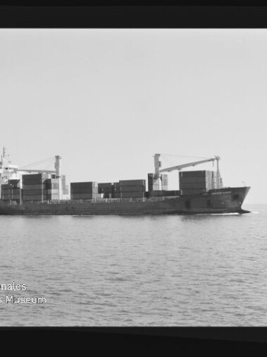Schwarzweißfoto eines Containerschiffs in Seitenansicht auf ruhiger See mit aufgestapelten Containern und zwei Ladekränen an Deck, im Hintergrund ein kleines Schiff und unten links das Wasserzeichen ‚Internationales Maritimes Museum Hamburg‘.