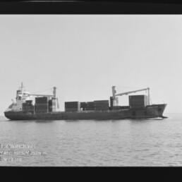 Schwarzweiß-Seitenansicht eines beladenen Containerschiffs mit aufgestapelten Containerblöcken und zwei Ladekränen an Deck, das ruhig auf dem Meer fährt; links unten sichtbar das Wasserzeichen des Internationalen Maritimen Museums Hamburg.