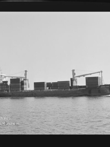 Schwarzweiß-Seitenansicht eines beladenen Containerschiffs mit aufgestapelten Containerblöcken und zwei Ladekränen an Deck, das ruhig auf dem Meer fährt; links unten sichtbar das Wasserzeichen des Internationalen Maritimen Museums Hamburg.