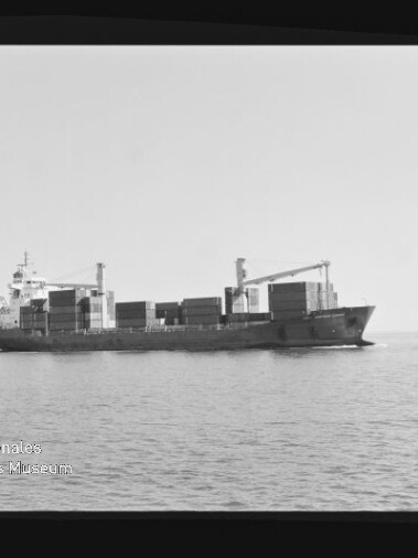 Containerfrachter mit aufgestapelten Ladecontainern und Bordkränen fährt in ruhiger See vor flachem Horizont; ein Vogel fliegt rechts am Himmel, unten links sichtbares Wasserzeichen des Internationalen Maritimen Museums Hamburg.