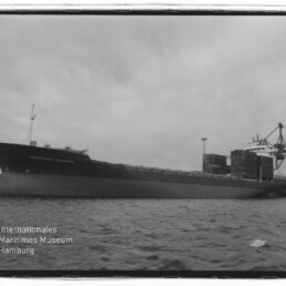 Schwarzweißaufnahme eines großen Containerschiffs in Seitenansicht am Hafen mit aufgetürmten Containern, Hafenkränen im Hintergrund, ruhiger Wasseroberfläche und dem Logo des Internationalen Maritimen Museums Hamburg in der Ecke.