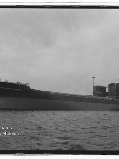 Schwarzweißaufnahme eines großen Containerschiffs in Seitenansicht am Hafen mit aufgetürmten Containern, Hafenkränen im Hintergrund, ruhiger Wasseroberfläche und dem Logo des Internationalen Maritimen Museums Hamburg in der Ecke.