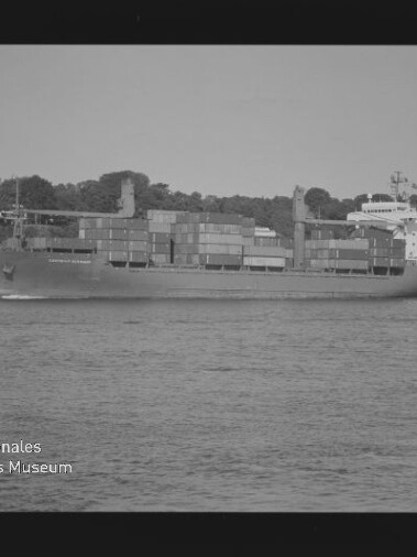Schwarzweißaufnahme eines beladenen Containerschiffs mit gestapelten Containern, das auf ruhigem Wasser an einer bewaldeten Uferlinie mit vereinzelten Häusern vorbeifährt; links unten sichtbar das Logo des Internationalen Maritimen Museums Hamburg und Filmrandbeschriftung.