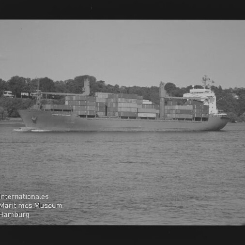 Schwarzweißaufnahme eines beladenen Containerschiffs mit gestapelten Containern, das auf ruhigem Wasser an einer bewaldeten Uferlinie mit vereinzelten Häusern vorbeifährt; links unten sichtbar das Logo des Internationalen Maritimen Museums Hamburg und Filmrandbeschriftung.