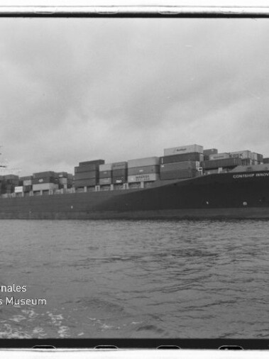 Großes Containerschiff mit zahlreichen aufgestapelten Frachtcontainern auf ruhigem Wasser unter einem bewölkten Himmel; links unten sichtbar das Wasserzeichen des Internationalen Maritimen Museums Hamburg.